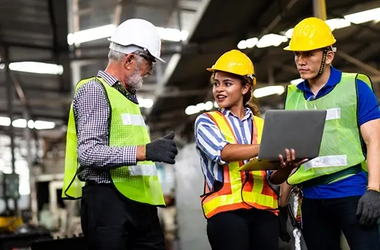 Diverse construction team wearing safety helmets and high visibility vests reviewing project data on laptop in warehouse Mobile