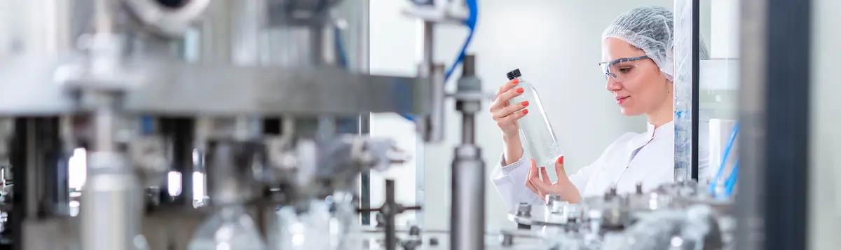 Female quality control technician in hairnet and safety glasses inspecting clear bottle on automated manufacturing line