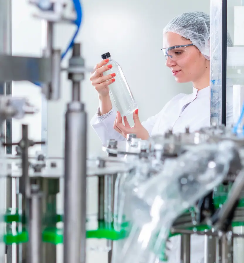 Female quality control technician in hairnet and safety glasses inspecting clear bottle on automated manufacturing line Mobile