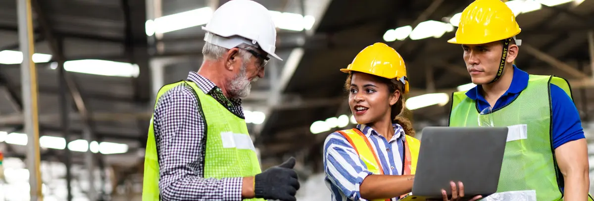 Diverse construction team wearing safety helmets and high visibility vests reviewing project data on laptop in warehouse